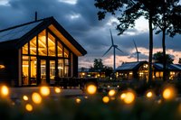 Modern eco-friendly resort cottages with warm glowing lights at dusk, wind turbines in background and illuminated string lights creating magical atmosphere in foreground.