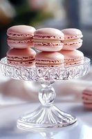 Pink macarons with delicate filling arranged on vintage crystal cake stand against soft white background, elegant dessert presentation for special occasions.
