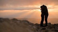 Silhouette of romantic couple embracing on mountain peak during golden sunset with dramatic cloudy sky and scenic landscape view for love and adventure concepts.