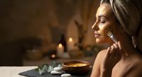 Young woman applying facial serum in candlelit spa setting with towel wrapped hair enjoying luxurious skincare routine and wellness treatment.