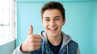 Happy teenage boy giving thumbs up gesture in casual denim jacket and hoodie against turquoise wall background for youth lifestyle and positive attitude concepts.