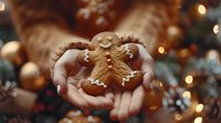 Hands holding freshly baked gingerbread man cookie with icing decoration against blurred Christmas lights and pine branches background, festive holiday mood.