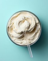 Creamy white frosting swirled in glass bowl with plastic spatula on mint blue background, overhead view showing smooth texture and soft peaks for baking and desserts.
