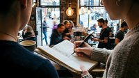 Musician reading sheet music at cafe counter with coffee cup, other patrons and staff visible in background, warm pendant lighting overhead.