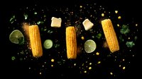 Fresh corn cobs with butter cubes, lime slices, and herbs floating on black background with scattered seasonings and dramatic lighting effect.