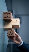 Businessman hand holding wooden cube with gear icon near stacked blocks showing business symbols for strategy planning and process management concepts.
