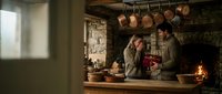 Medieval kitchen scene with two women in period clothing preparing food by stone fireplace with hanging copper pots and rustic wooden table setting.