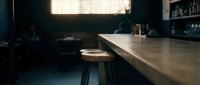 Modern bar counter with metal surface and wooden stool in dimly lit establishment, featuring shelves of bottles and warm window light for hospitality design.