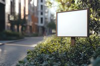 Blank white rectangular mockup sign board surrounded by green bushes on urban street with residential buildings and bokeh background. For advertising display.