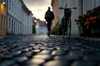 Atmospheric cobblestone street at dusk with silhouetted person walking and parked bicycle against historic buildings, orange flowers adding warmth to moody urban scene.