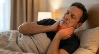 Middle-aged Caucasian man experiencing neck pain while lying in bed, holding his neck with hand, showing discomfort and muscle strain in bedroom setting.