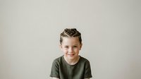 Young boy with curly brown hair, fair complexion, wearing olive green t-shirt, smiling at camera against neutral beige background. Suitable for family, education, and lifestyle marketing materials.