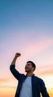 Young man with dark hair and beard, South Asian ethnicity, wearing navy button-up shirt over white t-shirt, raising fist in celebration against sunset sky. Success, achievement, motivation concept.