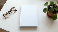 Minimalist workspace with blank notebook, eyeglasses and green plant on white desk. Clean office setup for productivity, planning and creative work.