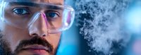 Bearded scientist wearing protective goggles observes chemical reaction with rising smoke in modern laboratory setting against blue background.