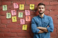 Caucasian man in denim shirt standing with crossed arms against red brick wall with colorful sticky notes showing question marks, representing doubt and decisions.