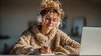 Young blonde woman in cozy sweater writing notes while listening to headphones at laptop, studying or working from home in warm comfortable setting.