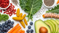 Colorful superfood ingredients arranged on white background including avocado slices, blueberries, kale leaves, goji berries, chia seeds and turmeric.