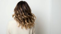 Woman with wavy brown hair and blonde highlights wearing white sweater from behind against neutral background for hair salon and beauty concepts.