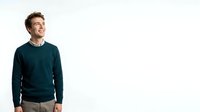 Young adult male with light skin tone and brown hair wearing dark teal crewneck sweater over collared shirt, smiling while looking to side against white background for portrait and lifestyle use.