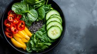 Fresh vegetable buddha bowl with spinach leaves, sliced bell peppers, cucumber, and black quinoa on dark plate against grey concrete background.