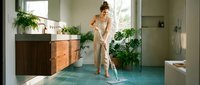 Woman in casual beige clothing cleaning modern bathroom floor with mop, surrounded by potted plants and wooden vanity, bright natural light from windows.