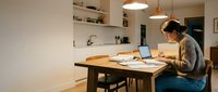 Young Asian woman working on laptop at wooden dining table in modern minimalist kitchen interior with pendant lighting and open shelving for remote work productivity.