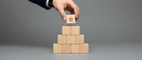 Businessperson placing wooden block on pyramid stack against gray background, representing growth, success, and strategic planning for corporate development.