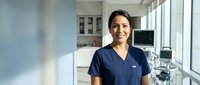 Smiling female healthcare professional in blue scrubs standing in modern medical office with dental equipment and bright windows creating welcoming atmosphere.