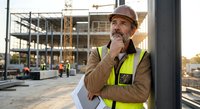 Thoughtful construction worker in safety vest and hard hat contemplating project at building site with concrete structure and crane in background.