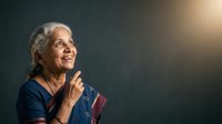 Senior woman with gray hair and South Asian ethnicity, wearing dark blue clothing, looking upward with hands clasped in prayer or gratitude gesture against dark background with warm lighting.