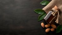 Natural almond oil bottle with fresh almonds and green leaves on dark stone background for skincare and aromatherapy wellness products.