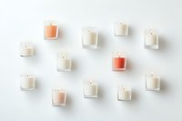Small glass candles in white and coral shades arranged on white background creating minimal composition with soft shadows and clean aesthetic.