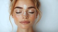Young woman with glowing skin and white dots decorative makeup on face against light background, eyes closed, natural beauty look and blonde hair in updo.