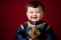 Joyful Asian baby wearing traditional blue and gold ceremonial outfit smiles brightly against deep red background, capturing cultural celebration and childhood innocence.