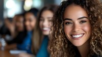Young mixed race woman with curly hair and bright smile in professional office setting with diverse coworkers in background, representing workplace diversity and inclusion.