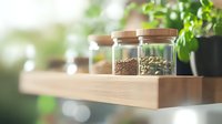 Glass jars with wooden lids containing coffee grounds and green coffee beans on wooden shelf, soft natural lighting with green plant bokeh background.