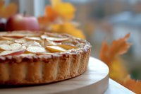 Rustic apple tart with sliced almonds on wooden serving board against autumn window background with blurred fall leaves creating cozy seasonal atmosphere.