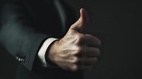 Close-up of businessman in formal suit showing thumbs up gesture against dark background, conveying approval, success, and positive feedback in corporate environment.