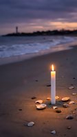Burning white candle on sandy beach at sunset with seashells and lighthouse silhouette in background creating peaceful memorial or meditation atmosphere.
