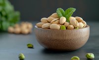 Fresh roasted pistachios in wooden bowl garnished with green leaves on dark background, selective focus with blurred foreground nuts for food photography.