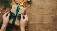 Hands wrapping Christmas gift with green ribbon on rustic wooden table with dried oranges and pine branches for holiday celebration.