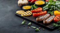 Grilled sausages with roasted vegetables and fresh herbs on dark wooden board. Rustic barbecue composition with colorful bell peppers, mushrooms and spices.