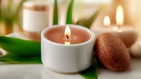 Burning aromatic candle in white ceramic holder with green bamboo leaves and brown stones creating zen atmosphere for spa and meditation practices.
