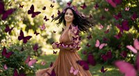 Young Asian woman in flowing brown dress dancing joyfully among purple butterflies in magical garden setting with warm sunlight and blooming flowers.