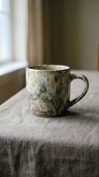 Rustic ceramic mug with weathered glaze sitting on linen fabric near window with natural light, suitable for home decor and lifestyle content.