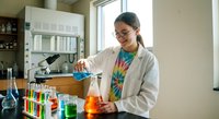 Young Hispanic female scientist in lab coat conducting chemical experiment with colorful solutions in modern laboratory setting for research and education.