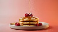 Stack of golden pancakes with fresh raspberries and syrup drizzle on white plate against warm pink coral gradient background for breakfast menu.