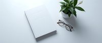 Minimalist workspace with closed white notebook, eyeglasses and green potted plant on clean desk surface for productivity and office organization.