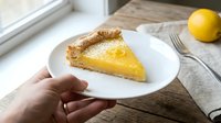 Slice of homemade lemon tart with golden custard filling and flaky pastry crust on white plate, hand holding dessert for food photography and culinary content.